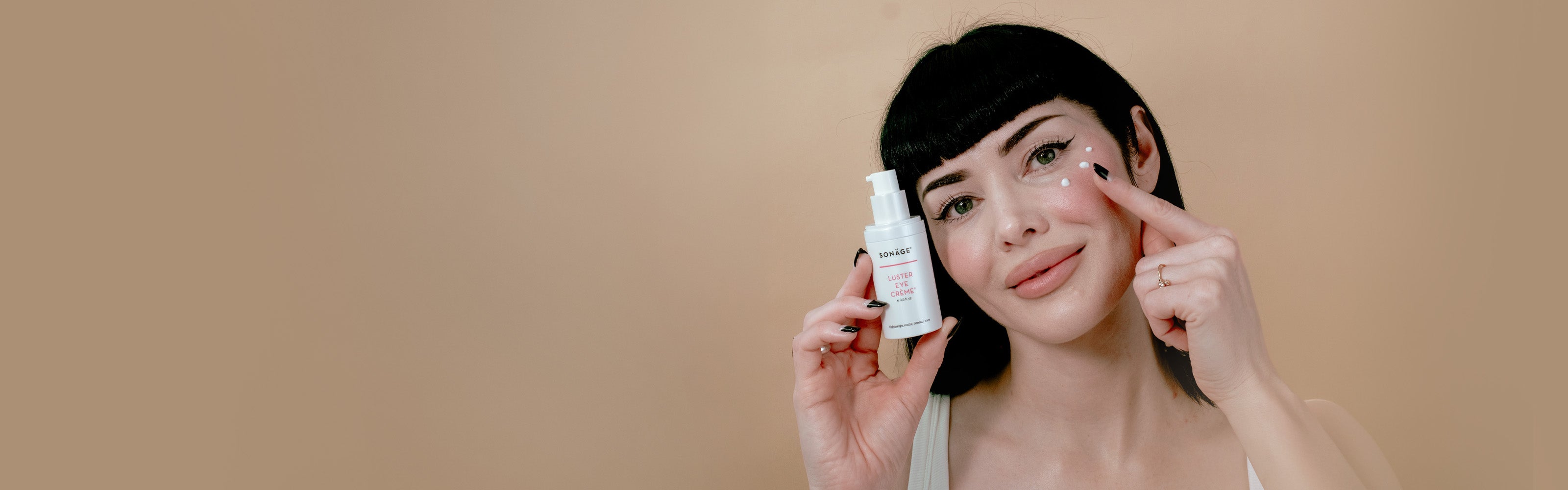 A woman with dark hair holds a luster eye cream in one hand while applying cream with her finger to her eye