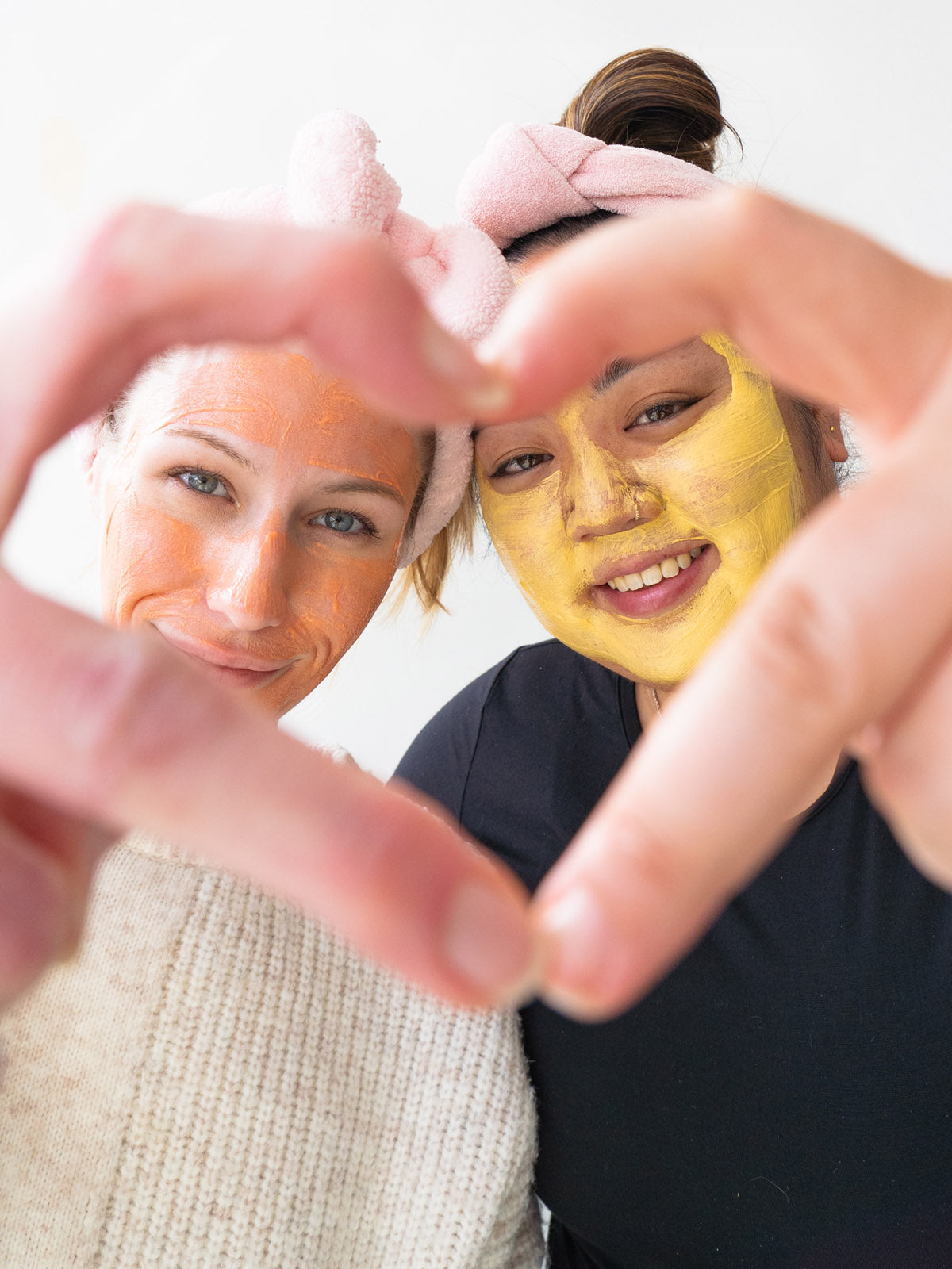 Two women, one with a yellow face mask, posing with their hands to form a heart shape.