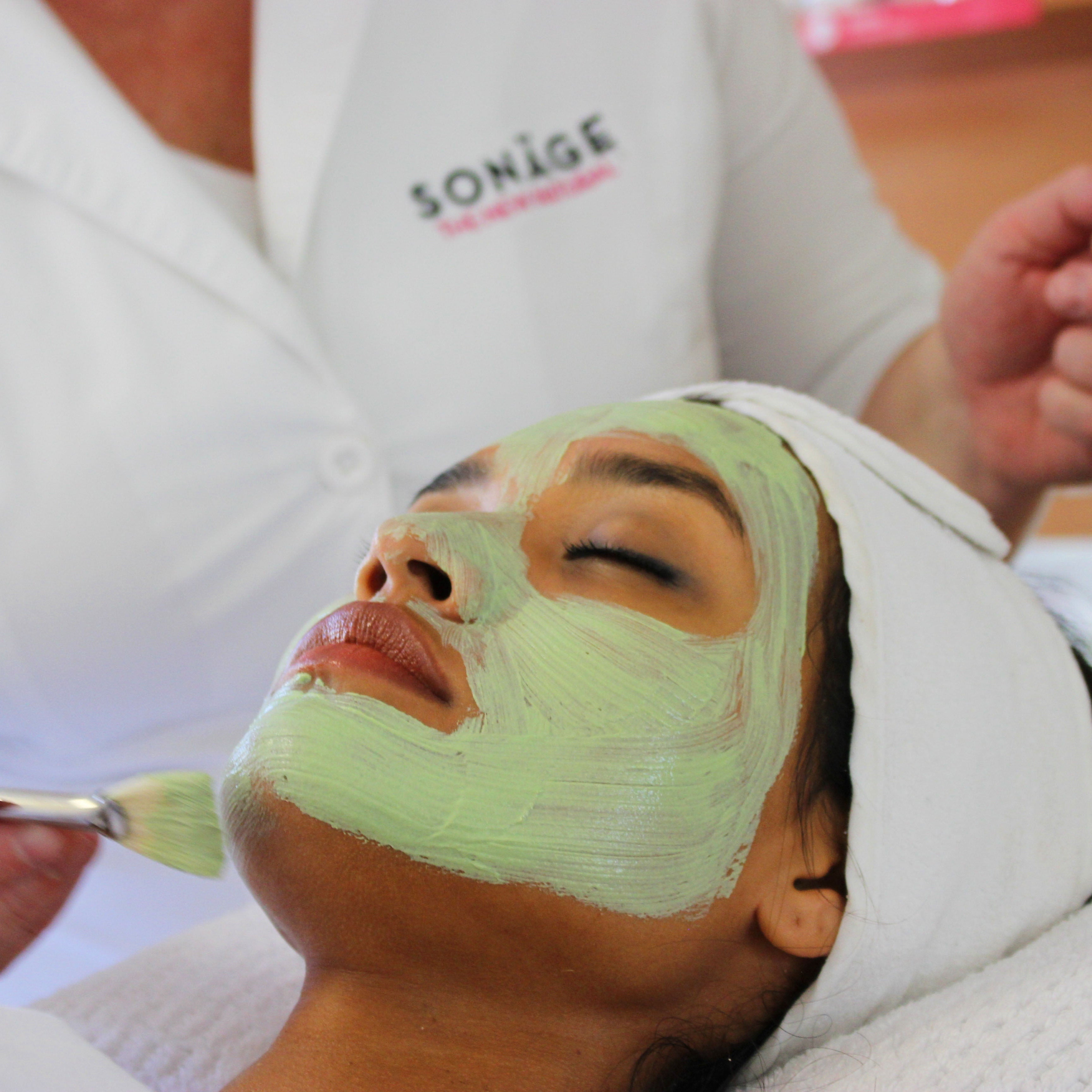 Person receiving a facial mask treatment with a green mask and brush, wearing a Sonage branded shirt.